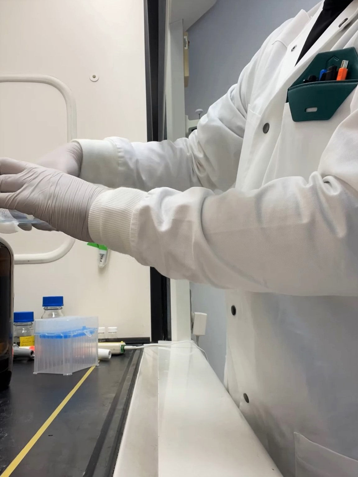 man in college chemistry teaching laboratory running experiments in fume hood while wearing white cotton lab coat with long knit cuffs safely covering his wrists while he reaches for chemistry glassware