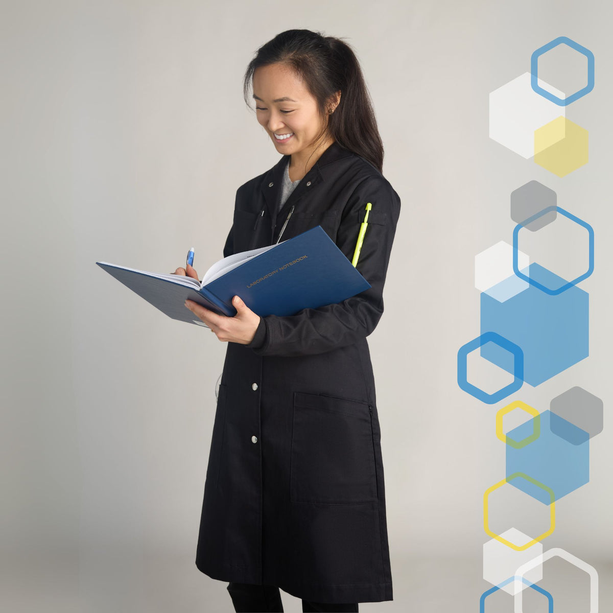 skinny short woman wearing a fitted long knee length black chemistry lab coat holding a science laboratory notebook and writing in it with a pen while holding utensils and markers in her pockets