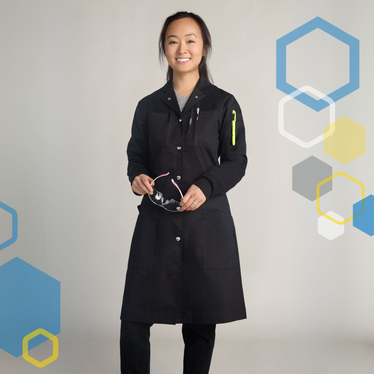 female scientist wearing women's microbiology lab coat in one hundred percent cotton black color, holding safety glasses with a marker in pockets on shoulder and metal snaps in front