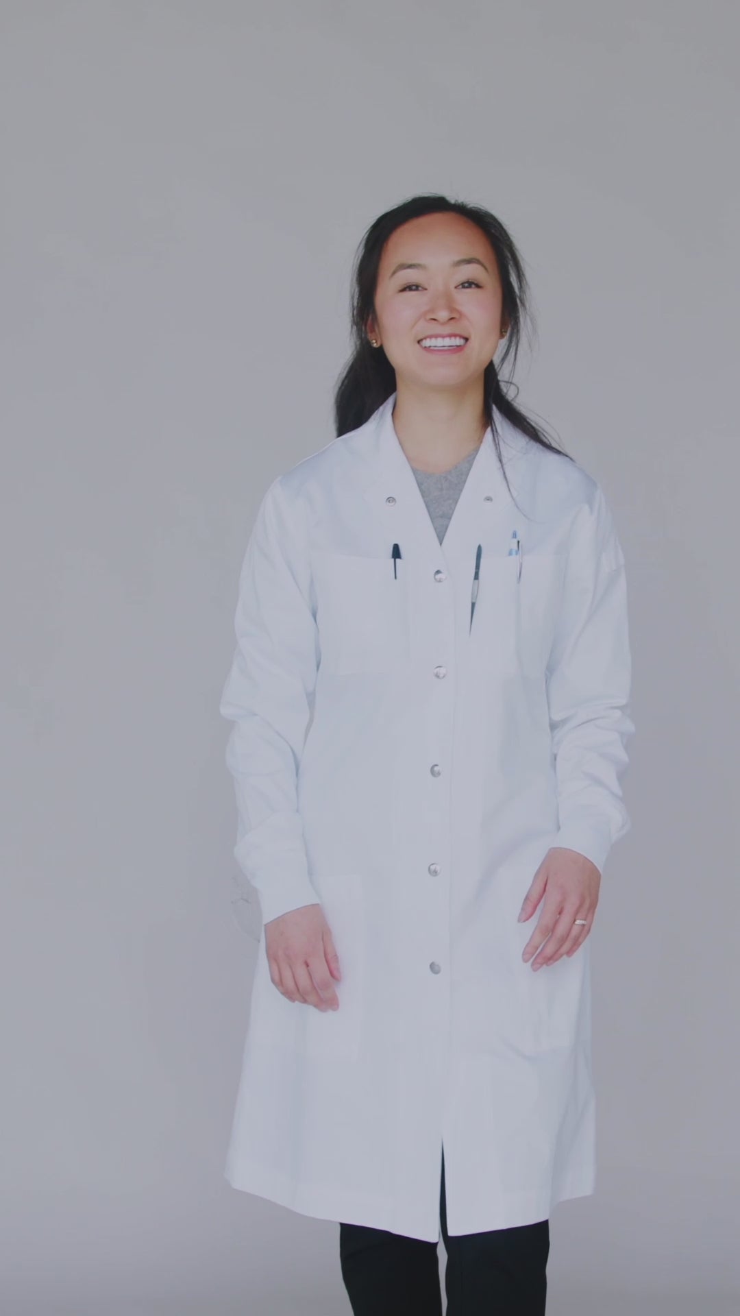 Petite woman wearing the XS black Curie lab coat showing pockets, zippers, snaps, knit cuffs, and adjustable belt