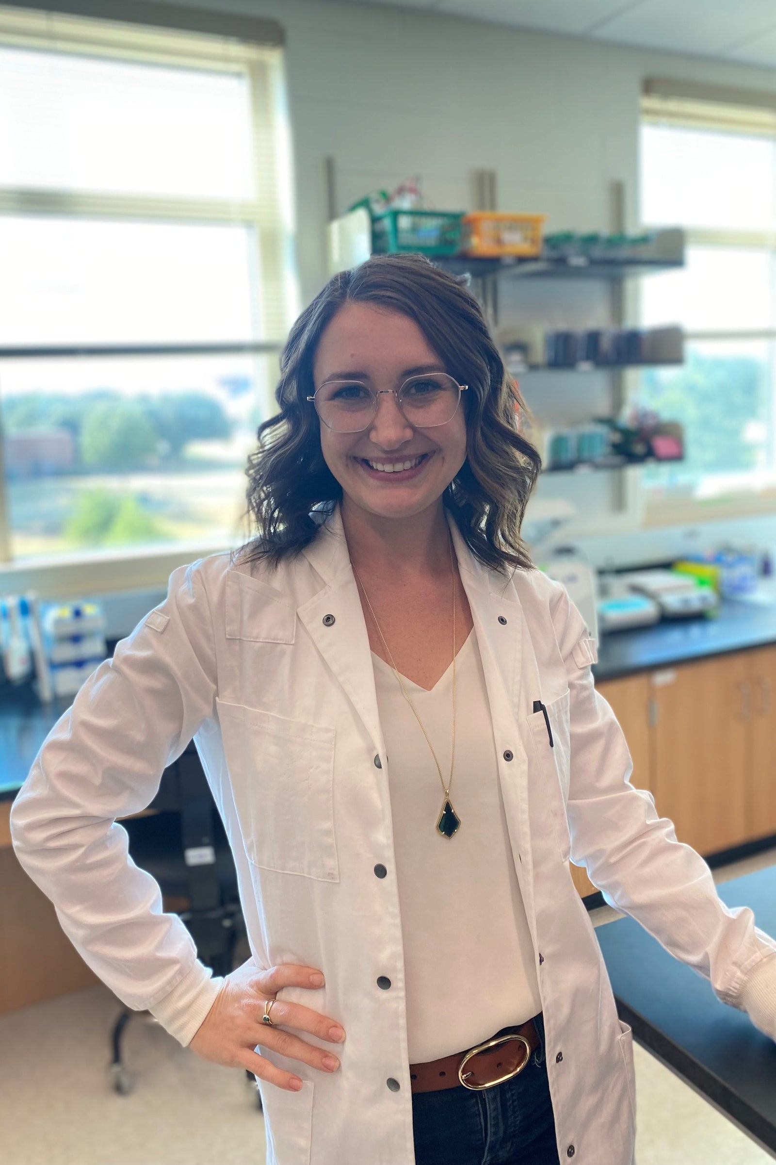 slender small woman wearing a well-fitted white one hundred percent cotton lab coat with metal snaps from the lab coat project testing. Standing in a microbiology laboratory with research equipment in the background.